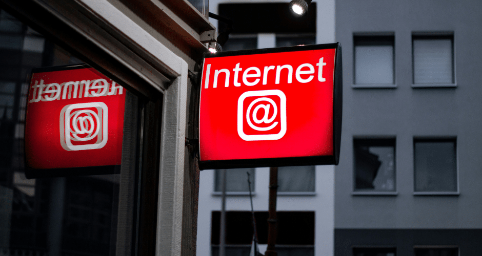 Uma placa vermelha com a palavra "Internet" e o símbolo @ está instalada em um prédio, refletindo em uma janela próxima - indicando a presença da Tributação sobre serviços de telecomunicações em meio ao cenário cinza de um apartamento ou escritório.