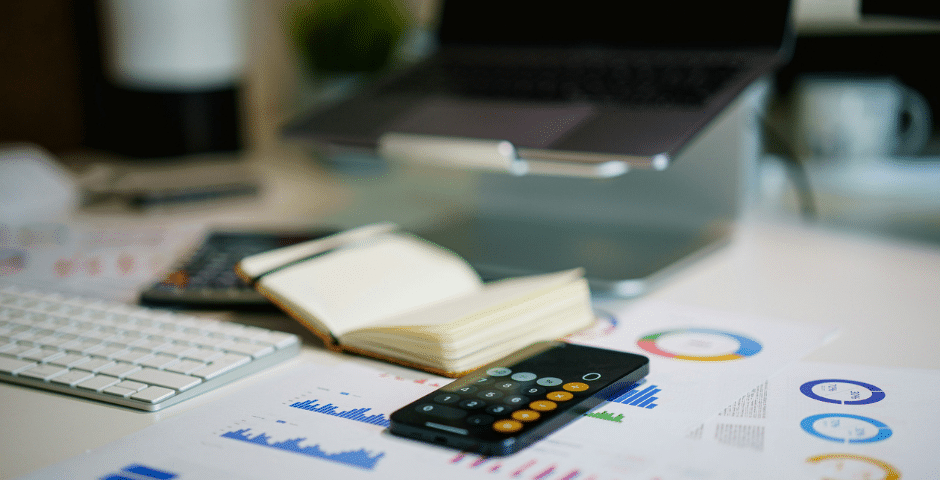 Um espaço de trabalho com um teclado, um smartphone exibindo um aplicativo de calculadora, um bloco de notas aberto, gráficos impressos com referência à EFD-Contribuições e um laptop em um suporte, sugerindo um ambiente de análise comercial ou financeira.