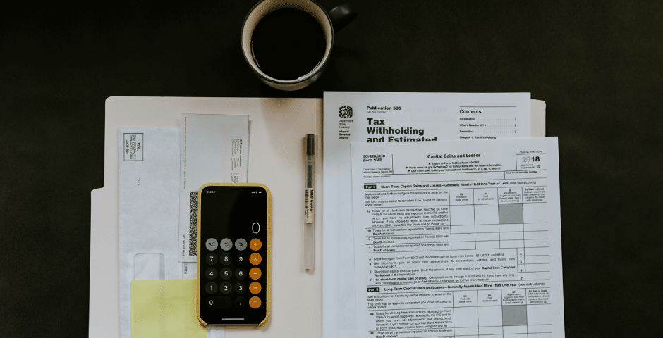 Uma escrivaninha com documentos fiscais, uma calculadora amarela, uma caneta, uma xícara de café, um envelope e uma pasta organizados ordenadamente, sugerindo que alguém está preparando ou declarando impostos diante dos novos impostos da reforma tributária.