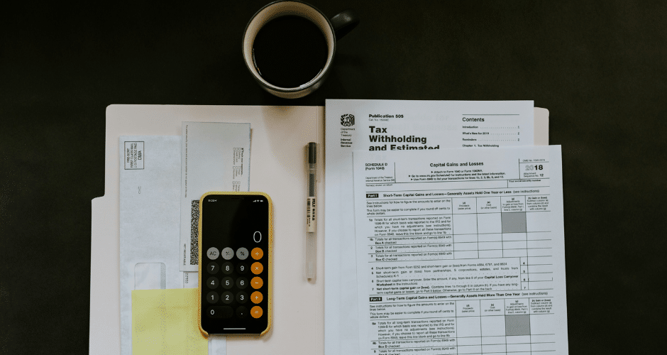 Uma escrivaninha com documentos fiscais, uma calculadora amarela, uma caneta, uma xícara de café, um envelope e uma pasta organizados ordenadamente, sugerindo que alguém está preparando ou declarando impostos diante dos novos impostos da reforma tributária.