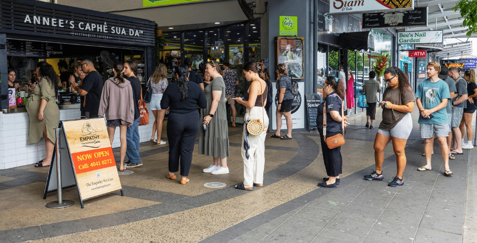 As pessoas ficam em uma longa fila do lado de fora do Annee's Caphe Sua Da, em uma calçada da cidade, algumas verificando seus telefones enquanto esperam. Nas proximidades, lojas vibrantes e placas anunciam serviços como emissão ilimitada de documentos fiscais.