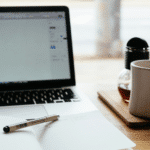 Um laptop, um caderno aberto com uma caneta, uma caneca e um pequeno vaso de flores sobre uma mesa de madeira, simbolizando o Fim do EPEC.