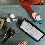 Uma pessoa segura um smartphone com um aplicativo de pagamento aberto perto de um tablet que exibe um catálogo de produtos em um balcão cinza, fazendo referência à Nota Técnica 2026.002, com pincéis, caixas de presente e produtos de beleza visíveis.