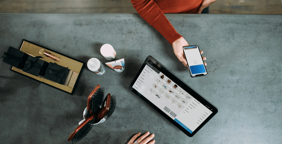 Uma pessoa segura um smartphone com um aplicativo de pagamento aberto perto de um tablet que exibe um catálogo de produtos em um balcão cinza, fazendo referência à Nota Técnica 2026.002, com pincéis, caixas de presente e produtos de beleza visíveis.