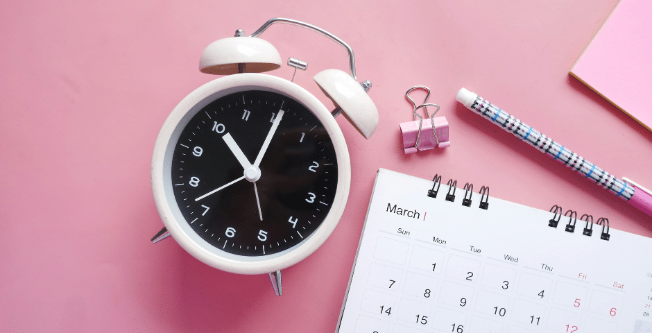 Um despertador branco, um calendário aberto para março, uma caneta, um clipe de fichário e um bloco de notas rosa estão dispostos em uma superfície rosa - perfeito para planejar prazos relacionados à NFS-e Nacional no Simples Nacional.