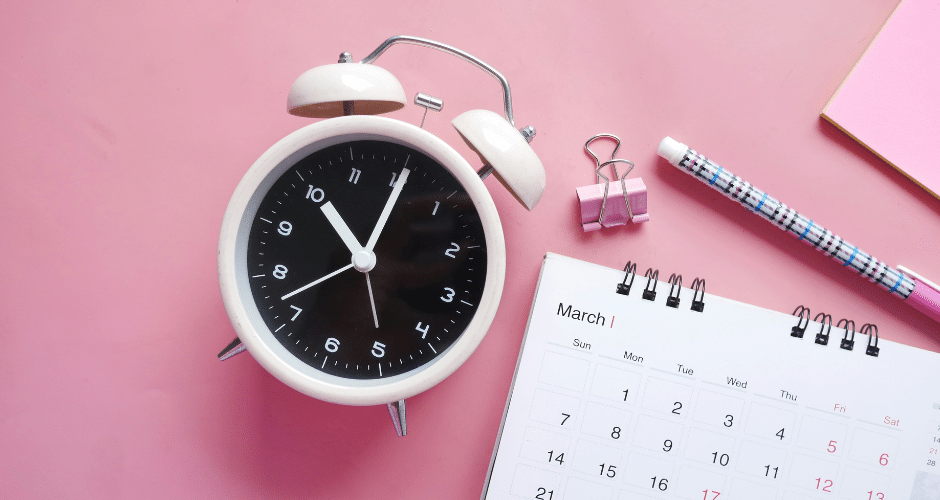 Um despertador branco, um calendário aberto para março, uma caneta, um clipe de fichário e um bloco de notas rosa estão dispostos em uma superfície rosa - perfeito para planejar prazos relacionados à NFS-e Nacional no Simples Nacional.