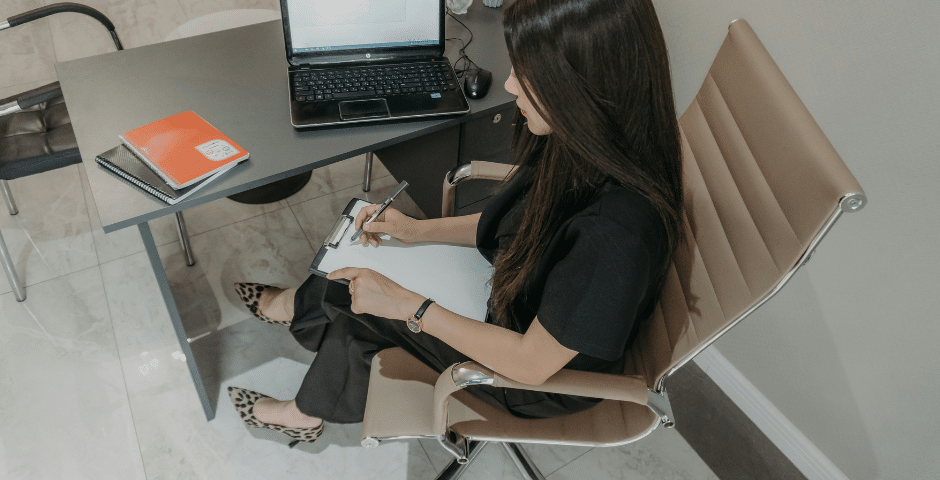 Uma mulher de cabelos longos e escuros está sentada em uma cadeira de escritório ao lado de uma mesa, escrevendo em uma prancheta. Sobre a mesa há um laptop, dois cadernos e documentos com a etiqueta NT 2026.002 Reforma Tributária CT-e. Ela veste roupas pretas e sapatos com estampa de leopardo.