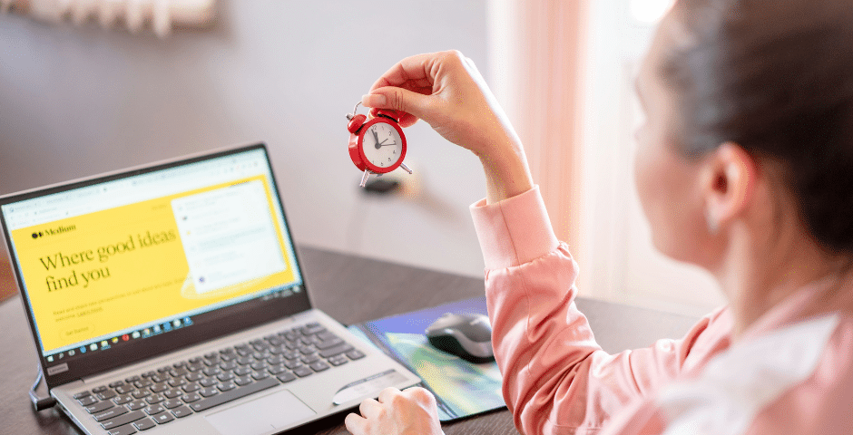 Uma mulher está sentada em uma mesa com um laptop, segurando um pequeno despertador vermelho. A tela do laptop exibe a frase “Onde as boas ideias te encontram”, possivelmente fazendo alusão a discussões sobre a obrigatoriedade do IBS e do CBS. Um mouse de computador está ao lado da mão dela.