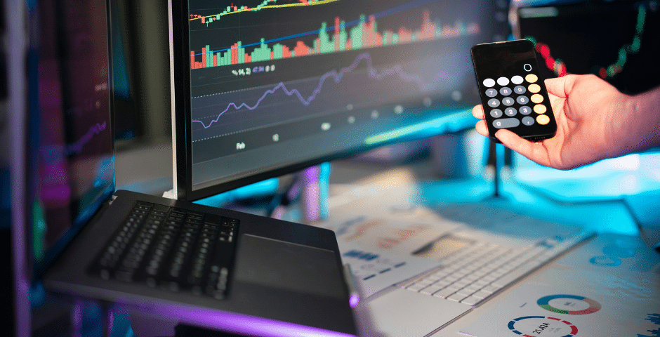 Uma pessoa segura um aplicativo de calculadora em um smartphone diante de telas de computador que exibem tabelas e gráficos financeiros coloridos, analisando os impactos da reforma tributária. Um laptop, um teclado e tabelas impressas também estão visíveis sobre a mesa.