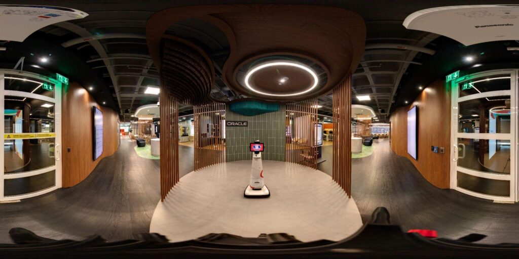 Uma vista de 360 graus do saguão de um escritório moderno, com um robô de telepresença no centro, detalhes em madeira, portas de vidro e a placa “Oracle” ao fundo — um espaço inspirador que incorpora perfeitamente o espírito de inovação da Comunidade Infinity do TecnoUpdate.