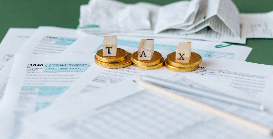 Moedas de ouro com letras de madeira que formam a palavra “TAX” estão dispostas sobre formulários fiscais espalhados, recibos e um lápis — simbolizando a preparação da declaração de imposto de renda com a burocracia semelhante à descrita no Manual do Contribuinte GNR-e.