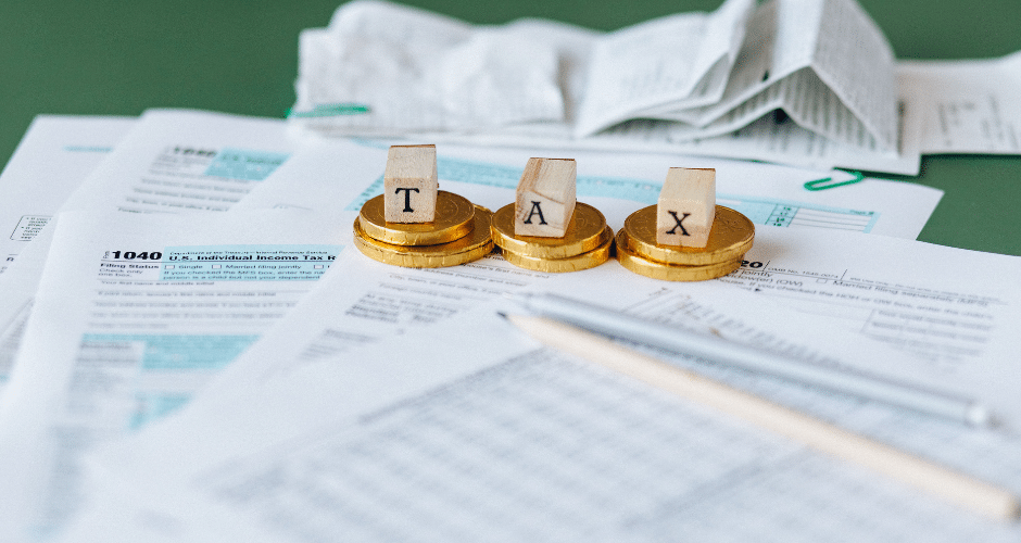Moedas de ouro com letras de madeira que formam a palavra “TAX” estão dispostas sobre formulários fiscais espalhados, recibos e um lápis — simbolizando a preparação da declaração de imposto de renda com a burocracia semelhante à descrita no Manual do Contribuinte GNR-e.