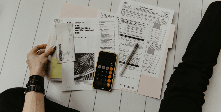 Uma pessoa está sentada no chão com documentos fiscais, uma caneta, a calculadora do smartphone e envelopes espalhados ao redor, aparentemente organizando ou preparando a declaração de impostos de acordo com a Tributação Regular do IBS e do CBS.
