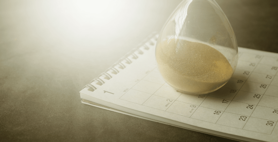 Uma ampulheta com areia escorrendo está colocada sobre um calendário aberto, simbolizando a passagem do tempo e os prazos, um lembrete visual da importância de acompanhar o cronograma dos documentos fiscais.