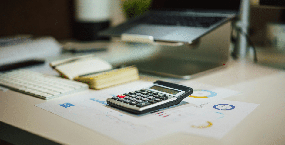 Uma calculadora sobre uma mesa, com gráficos financeiros, um caderno aberto, um teclado e um laptop ao fundo, ilustrando o processo de retenção de impostos na nota fiscal durante a análise financeira.