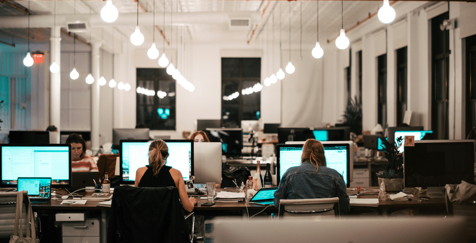 Um escritório em plano aberto, com pessoas trabalhando até tarde em suas mesas, iluminadas por lâmpadas suspensas. As telas dos computadores brilham enquanto os funcionários se concentram em tarefas como a finalização de relatórios sobre a Nota Técnica EFD-Reinf 04/2026 em um ambiente de trabalho moderno.