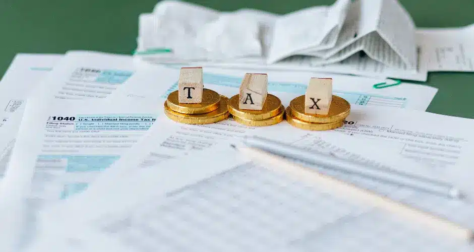 Pilhas de moedas com blocos de madeira que formam a palavra “TAX” estão sobre vários formulários fiscais e documentos financeiros, ao lado de um exemplar do manual do contribuinte GNR-e, com recibos e uma prancheta ao fundo.