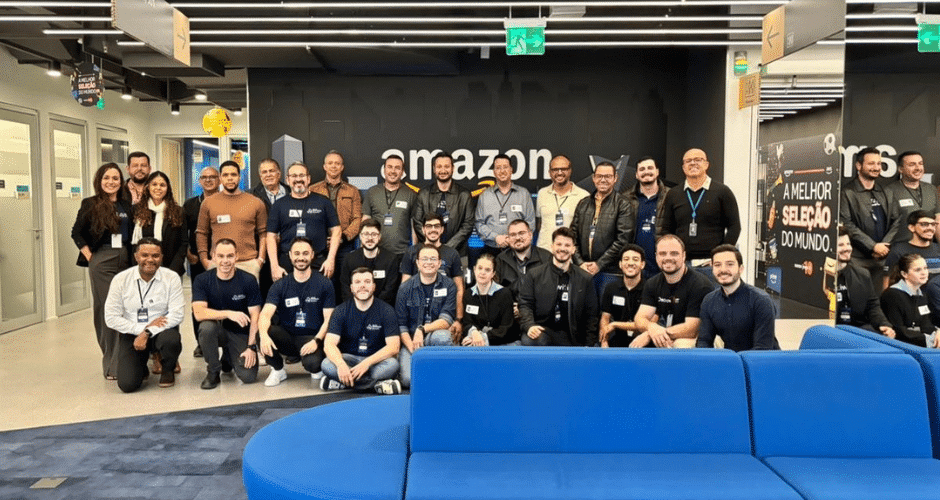 Um grande grupo de pessoas posa e sorri para uma foto em frente a uma placa da Amazon, dentro de um escritório com decoração moderna e sofás azuis, celebrando seu compromisso comum com a excelência na experiência do cliente.