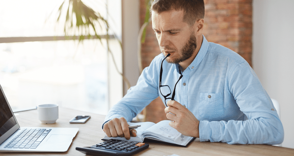 Um homem vestindo uma camisa azul-clara está sentado à mesa com um laptop, um caderno e uma calculadora; ele segura os óculos e olha pensativo para o caderno, talvez analisando dados relacionados ao FCP (Fundo de Combate à Pobreza).