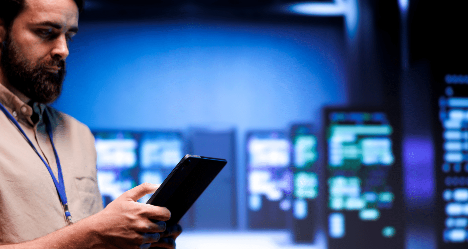 Um homem barbudo usa um tablet em uma sala de servidores mal iluminada, cercado por fileiras de racks de servidores iluminados ao fundo, enquanto monitora a conformidade dos dados de acordo com o Regime Especial.